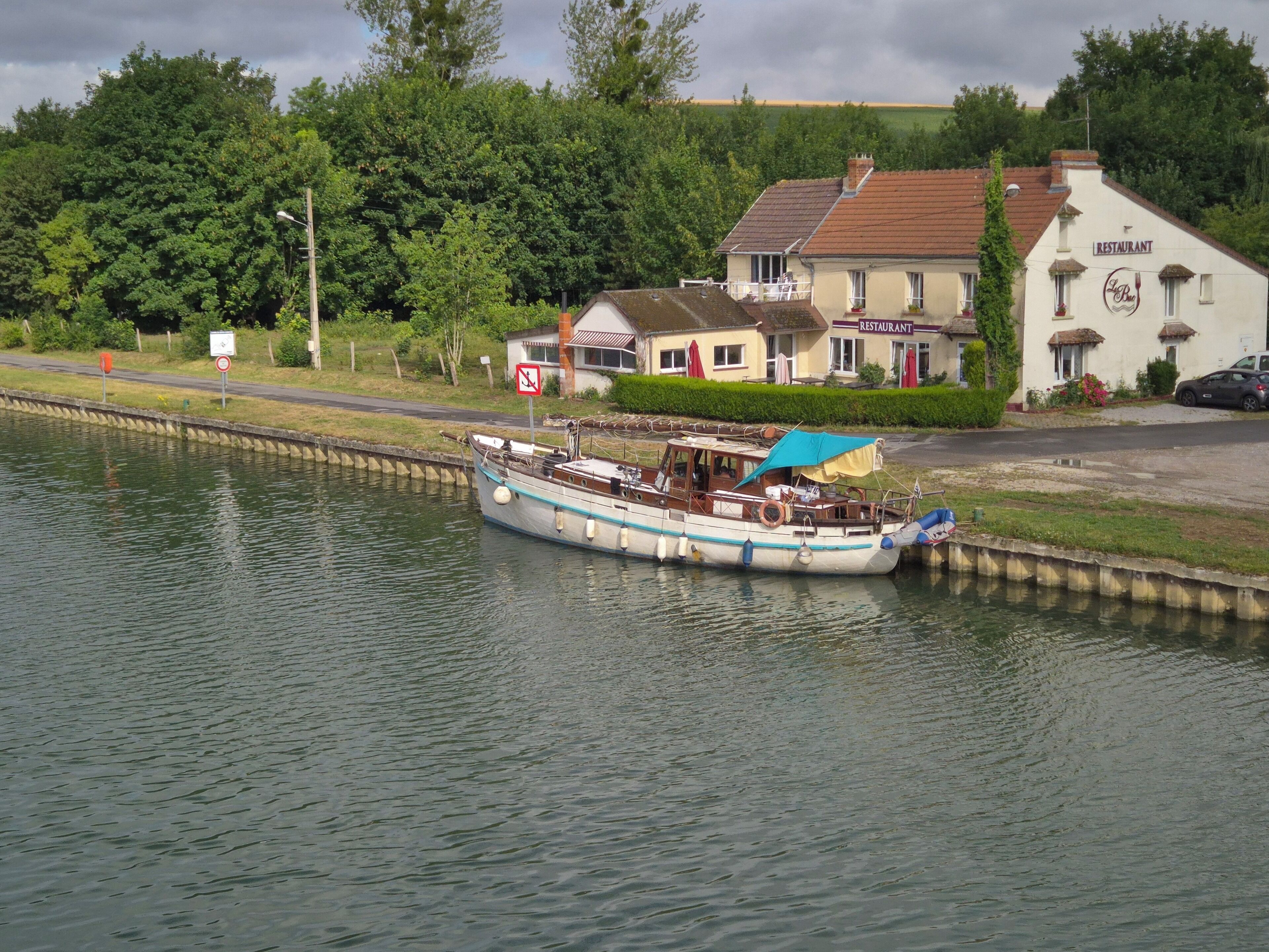 Een smalboot afgemeerd aan een kanaal, met een restaurant en woongebouwen op de achtergrond.