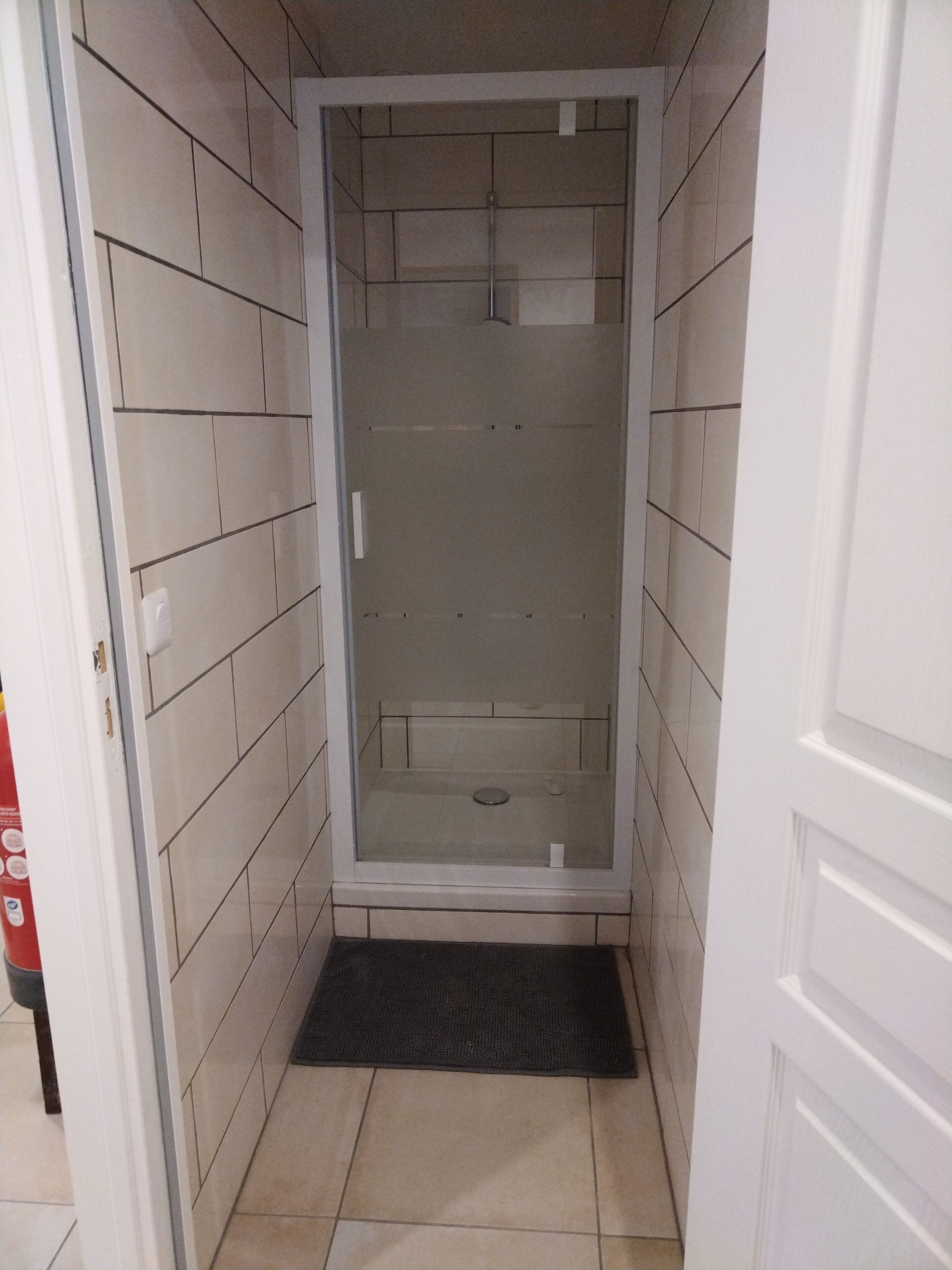 A small, enclosed shower area with tiled walls and floor, a glass door, and a black mat at the entrance.