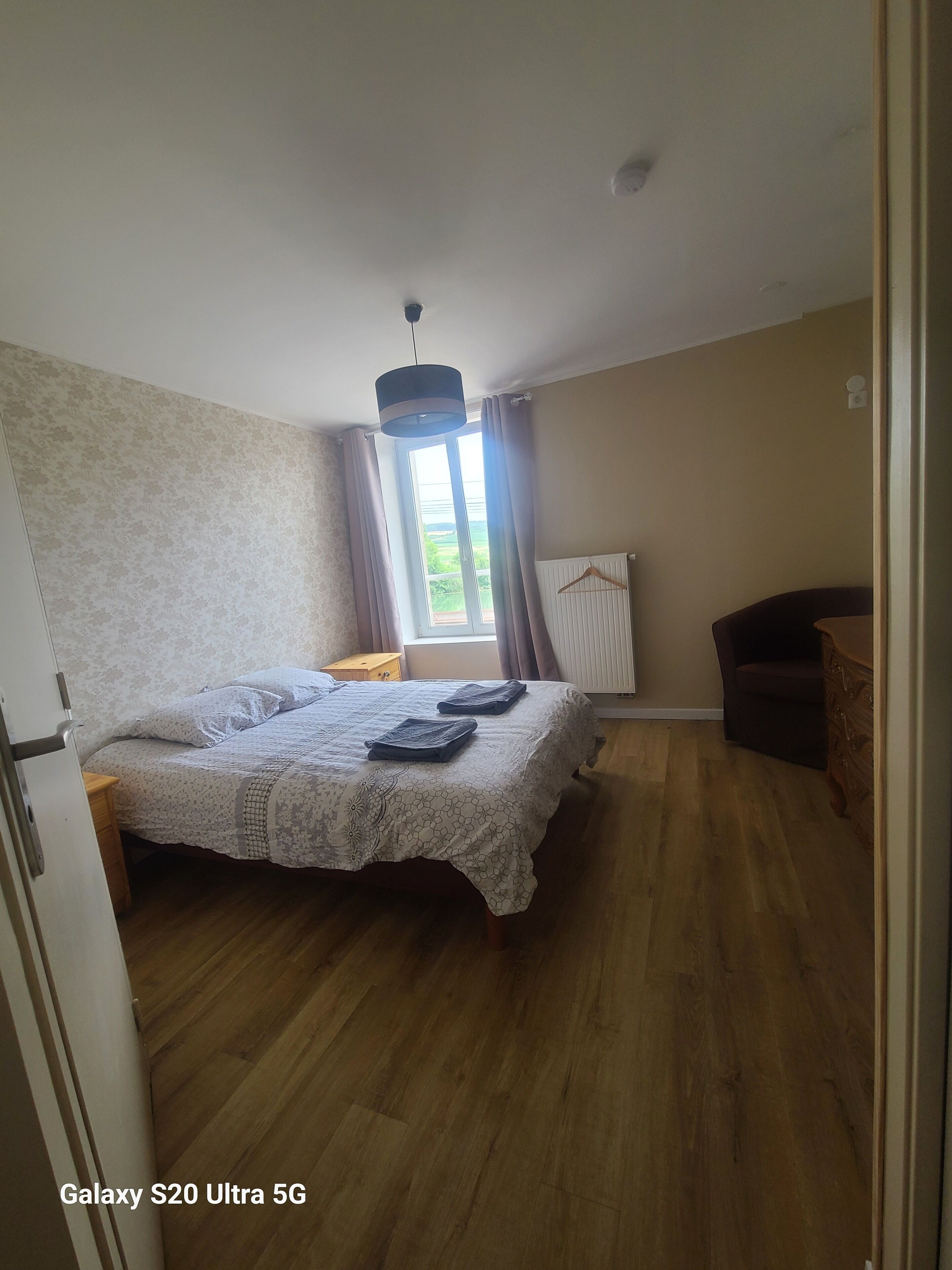 A bedroom with a bed, a wooden floor, a window with curtains, a radiator, and a hanging light fixture.