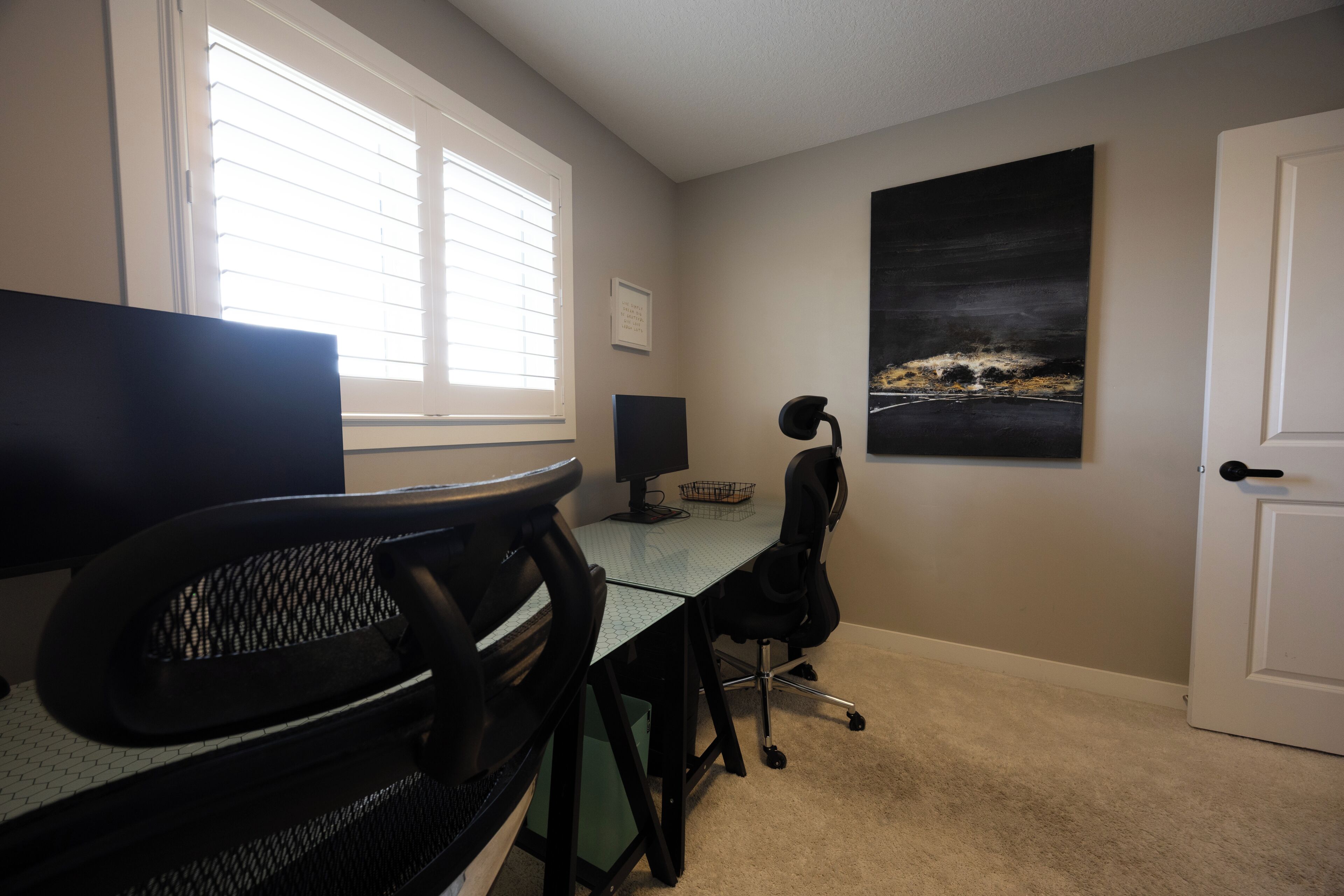A room with a desk, computer, chair, window, artwork, and door.