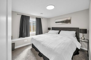 A bedroom with a bed, a window with curtains, a bedside table with a lamp, and a wall-mounted picture.