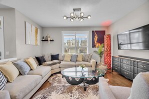 A living room with a sectional sofa, a glass coffee table, a TV, and a decorative wall piece.