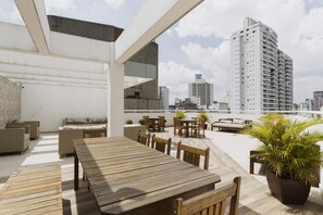 An outdoor terrace with a long wooden table and chairs, surrounded by potted plants and a city skyline view. - Av0203 Studio Avanhandava (São Paulo)