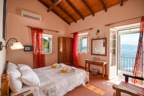 A bedroom with a bed, a wooden wardrobe, a bedside table, a mirror, and a view of the sea through an open door.