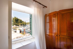 A room with a wooden wardrobe, a window with white curtains, and a view of a marina with boats and a forested hill.