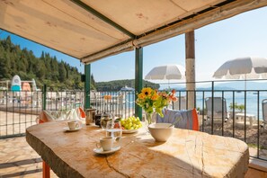 Et bord med en kop, et glas vand, en skål druer og en vase med blomster på en terrasse med udsigt over en strand og havet.