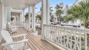 A wooden porch with white railings, a covered seating area, and a view of neighboring houses and palm trees.