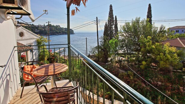 A balcony with a table and chairs, overlooking a coastal area with trees and a view of the sea. - Zoulis Andonis in Kalami (Corfu)