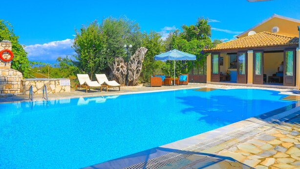 Une piscine d’un bleu éclatant, dotée de chaises longues, un mur de pierre et un bâtiment surmonté d’un toit en tuiles.