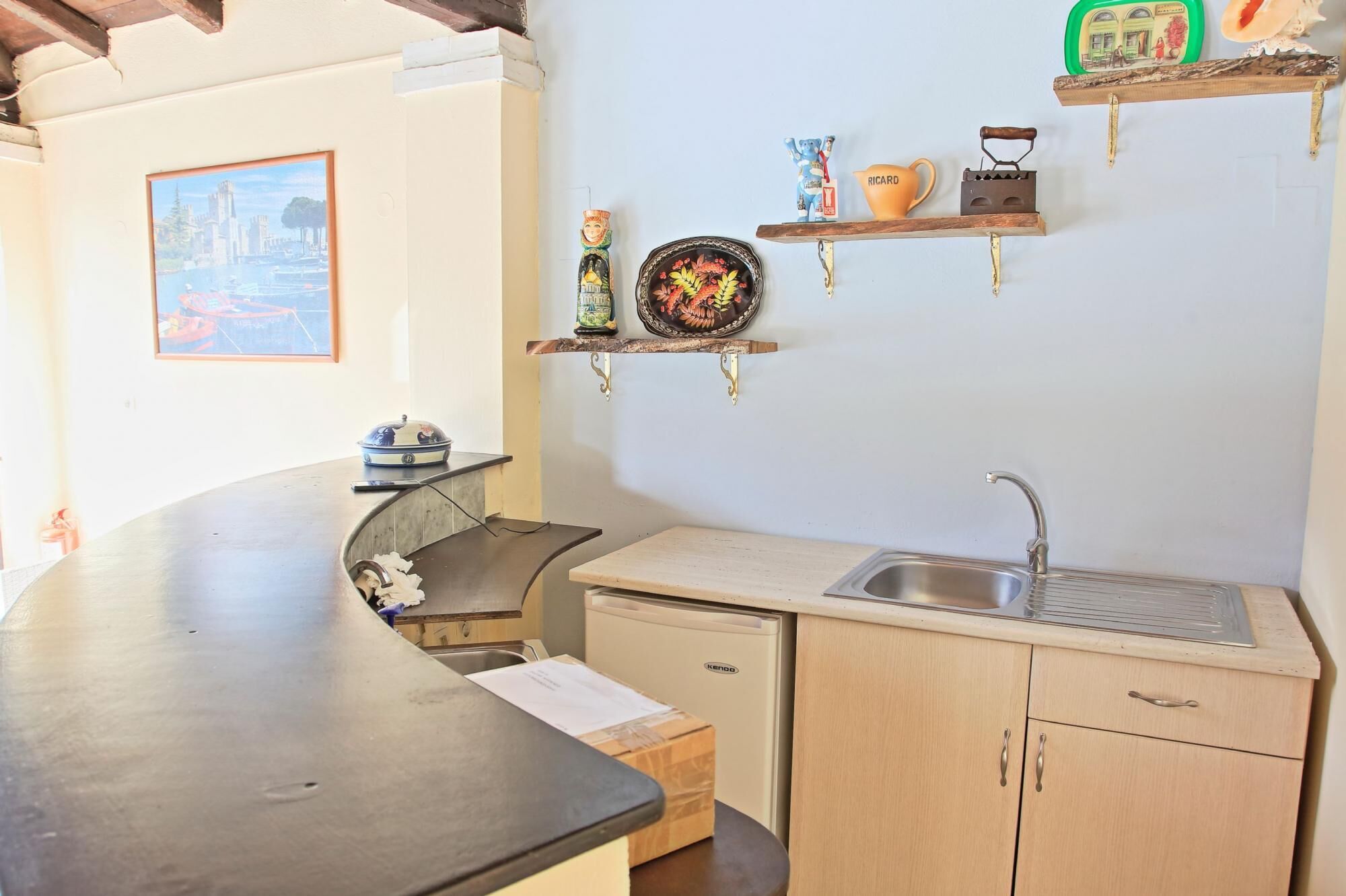 Uma cozinha compacta com pia, um frigorífico pequeno e bancada. Existem objetos decorativos nas prateleiras acima da bancada.