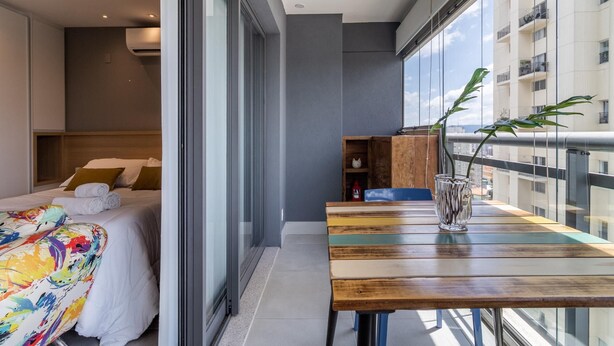 A modern hotel room with a bed, a wooden table, and a view of the city through large windows.