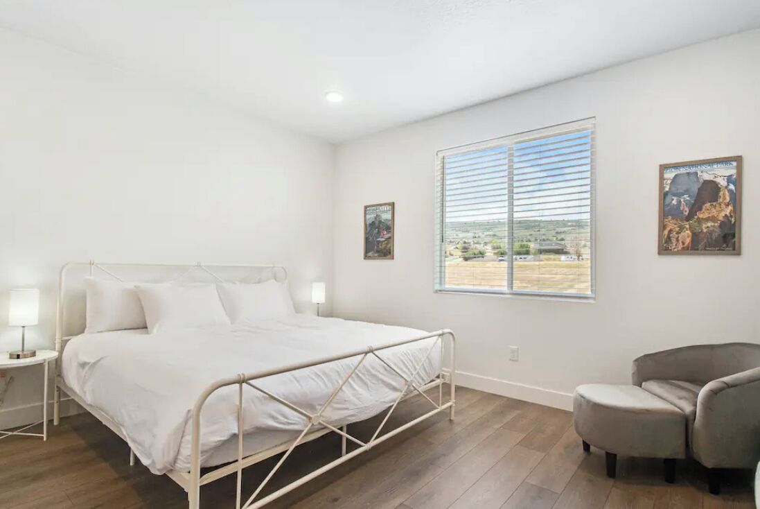 A bedroom with a metal bed frame, a single bedside table with a lamp, a chair, and two windows with blinds.