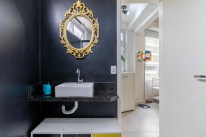 A modern bathroom with a black wall, a gold-framed mirror, a white sink, and a blue bottle on the counter. - Av0606 Studio Avanhandava (São Paulo)