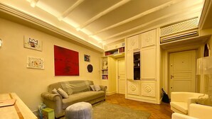 Un salon comprenant un canapé, un pouf et une table basse. On y trouve également une bibliothèque et une porte donnant sur une autre pièce.
