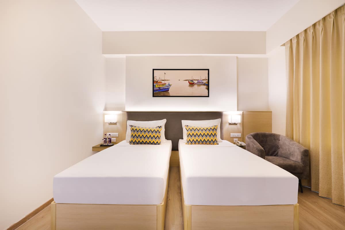 A hotel room with two single beds, a headboard with a framed picture, bedside lamps, a chair, and a curtain.