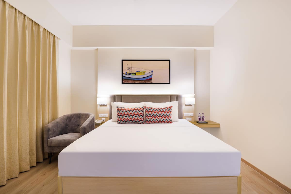 A hotel room with a bed, a chair, a nightstand, a framed picture on the wall, and a window with curtains.