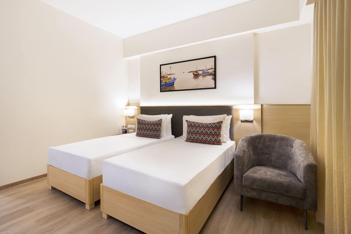 A hotel room with two beds, a grey armchair, a wall-mounted picture of boats, and a light-colored wooden floor.