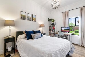 A bedroom with a bed, bedside tables, a desk, a chair, a bookshelf, and a window with a view of greenery.