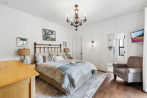 Un dormitorio con cama, mesitas de noche, una silla, un candelabro, un televisor y una puerta que da a un baño.