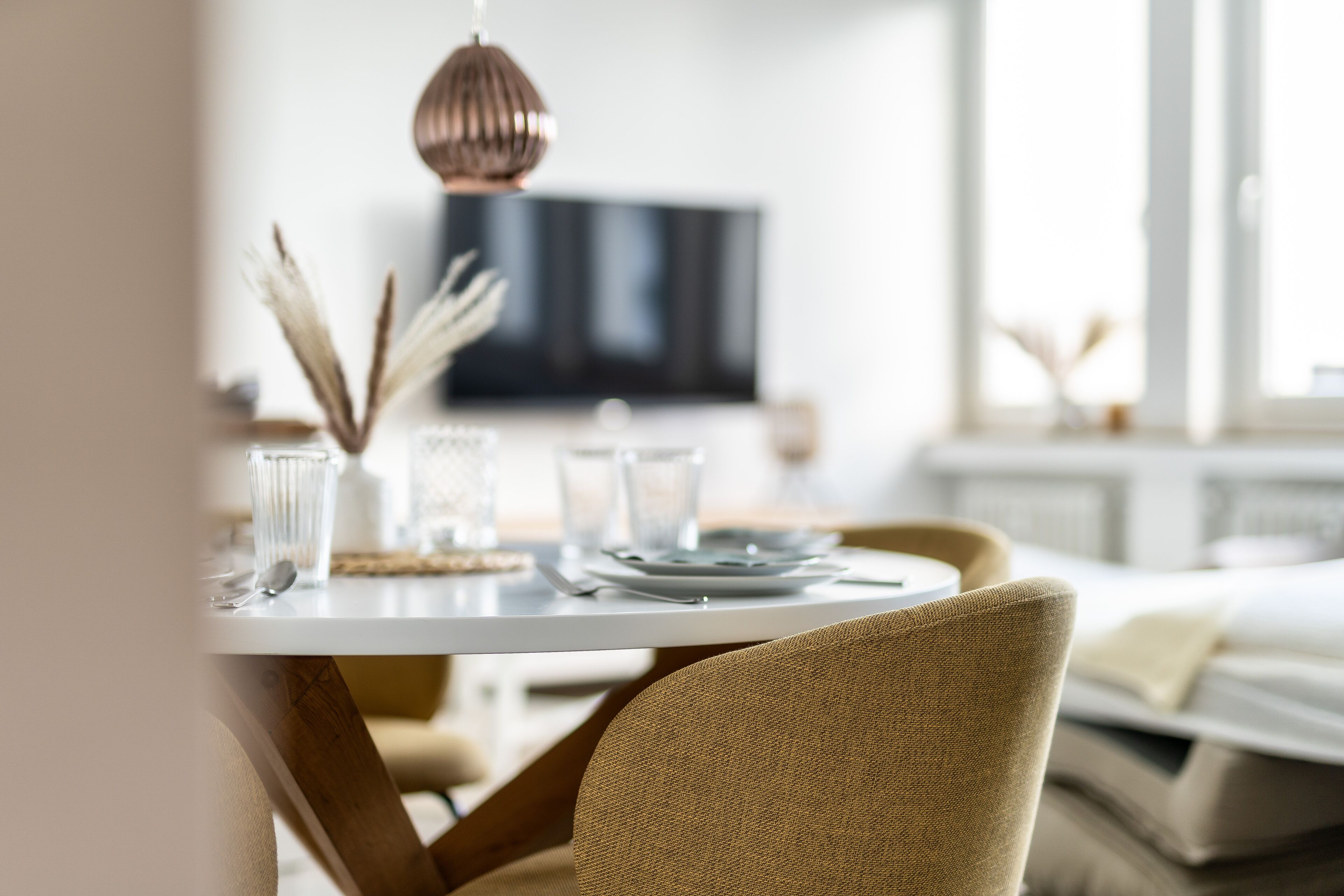 Ein Esstisch mit weißer Tischdecke, eine Pendelleuchte aus Kupfer und ein Fernseher im Hintergrund.