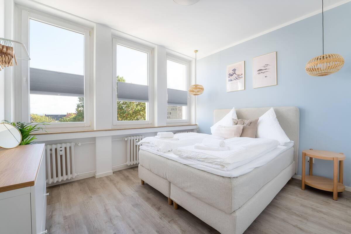 Quarto com cama, janelas amplas, radiador e uma pequena mesa de cabeceira de madeira.