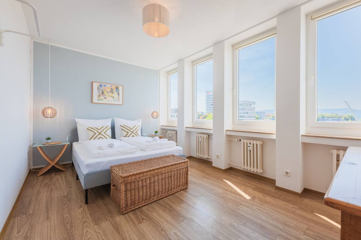 Quarto moderno com uma cama grande, um banquinho de vime, uma mesa de cabeceira e vista da cidade através de amplas janelas.