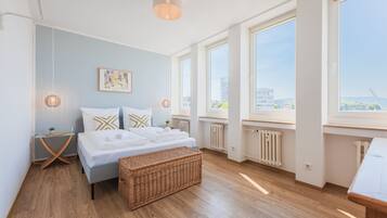 A modern bedroom with a large bed, a wicker ottoman, a bedside table, and a view of the city through large windows.