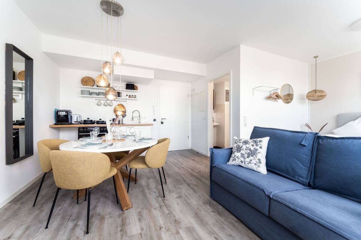 Uma sala de estar moderna com um sofá azul, uma mesa de jantar com cadeiras e uma área de cozinha ao fundo.