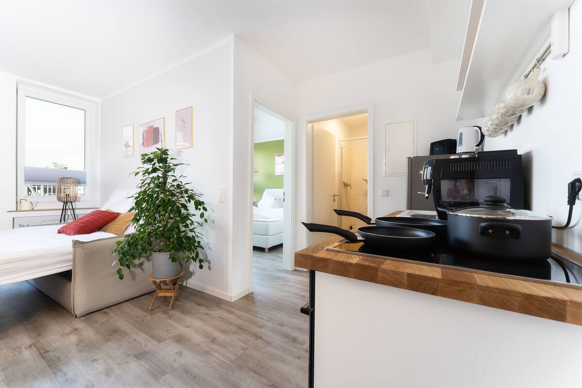 Uma cozinha moderna com forno embutido e máquina de café, uma sala de estar aconchegante com sofá e planta, e um quarto iluminado com cama e janela.