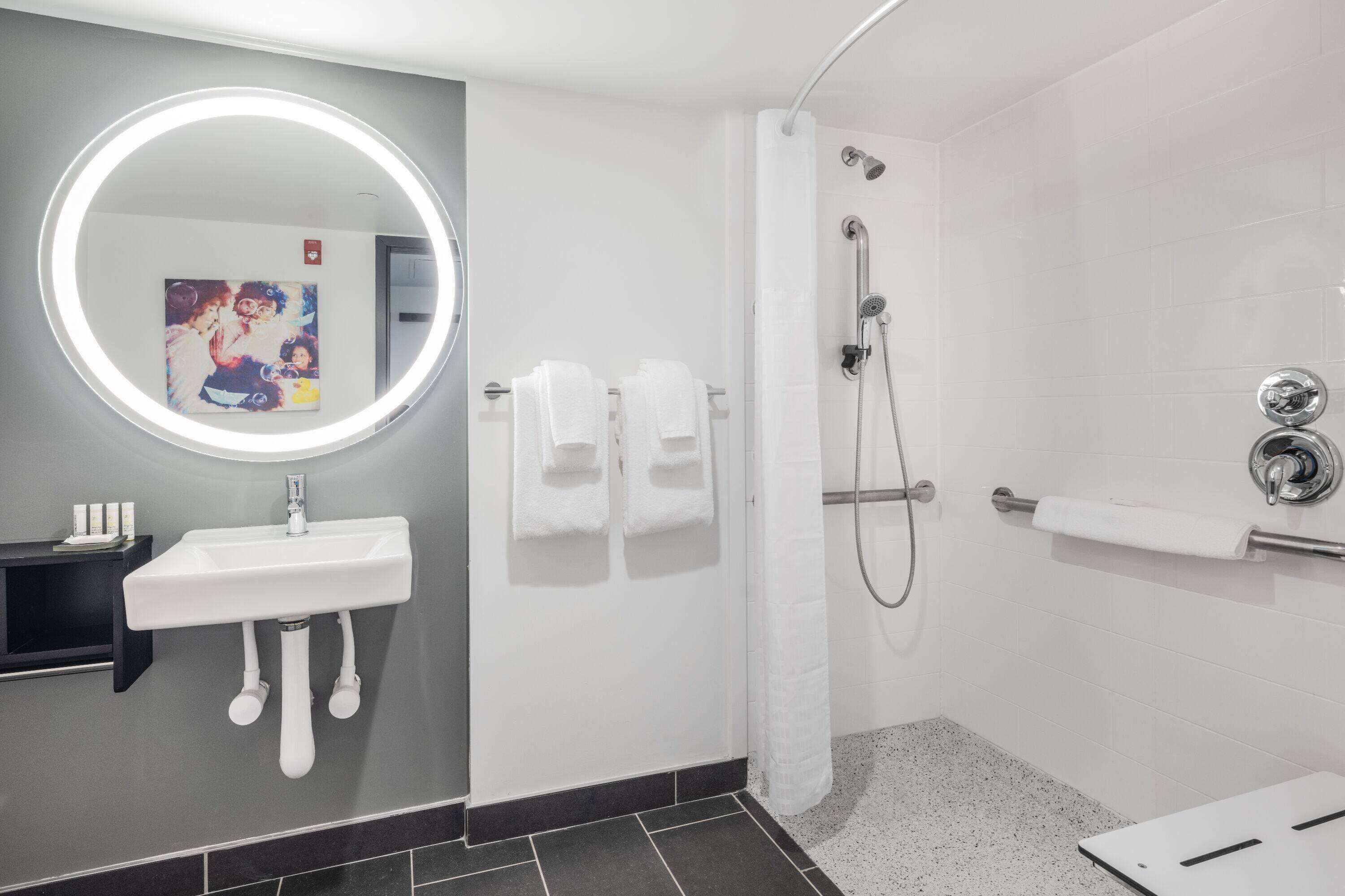 A modern bathroom with a white sink, a round mirror, a shower, and a towel rack.