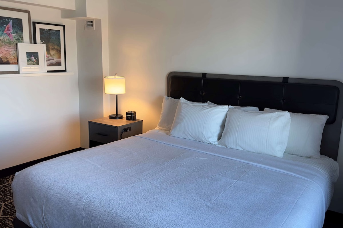 Ein ordentlich gemachtes Bett mit weißer Bettwäsche, ein Nachttisch mit Lampe und gerahmte Bilder an der Wand.