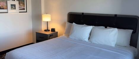 A neatly made bed with white linens, a bedside table with a lamp, and framed pictures on the wall.