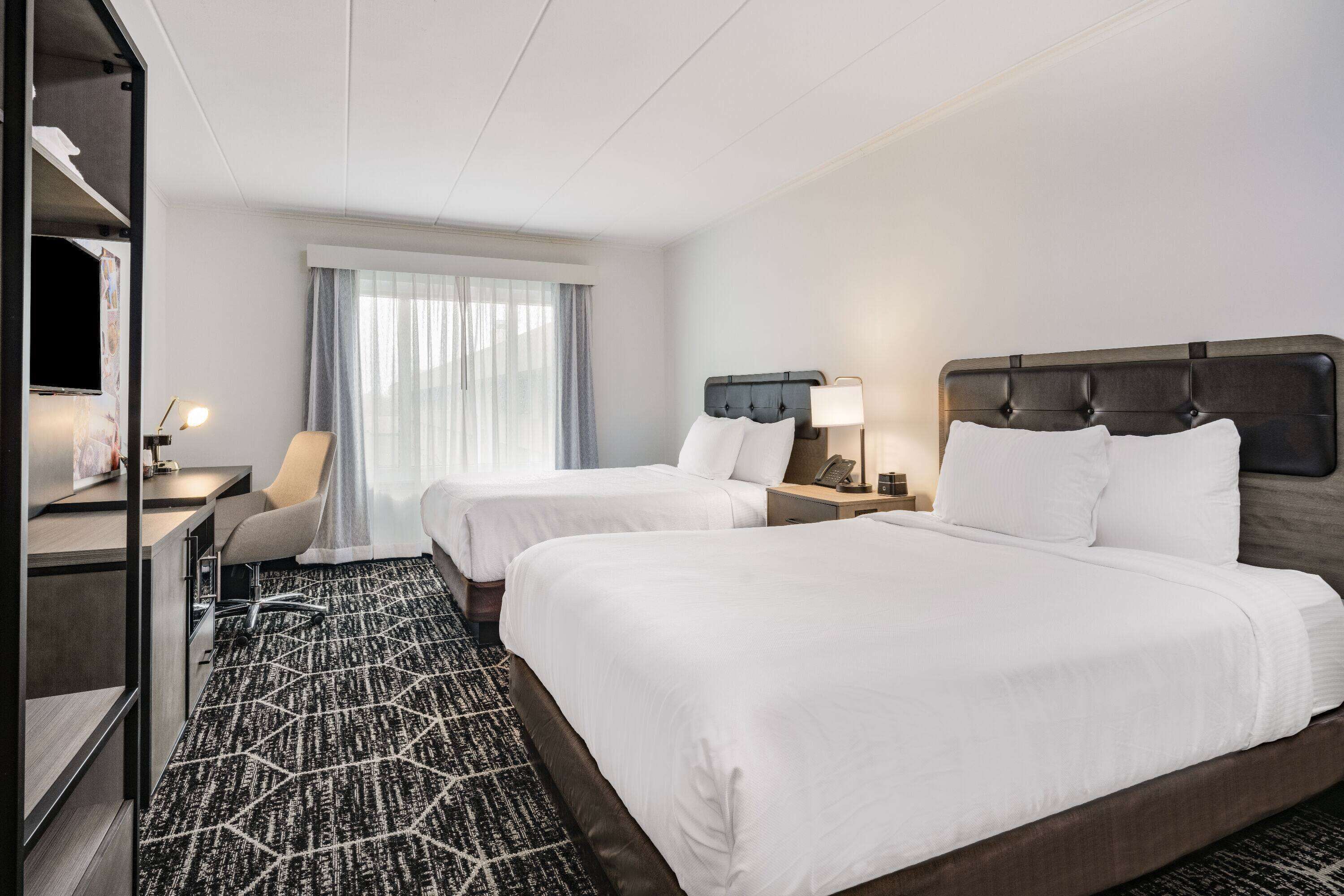 A hotel room with two beds, a desk, a chair, and a large window with curtains.