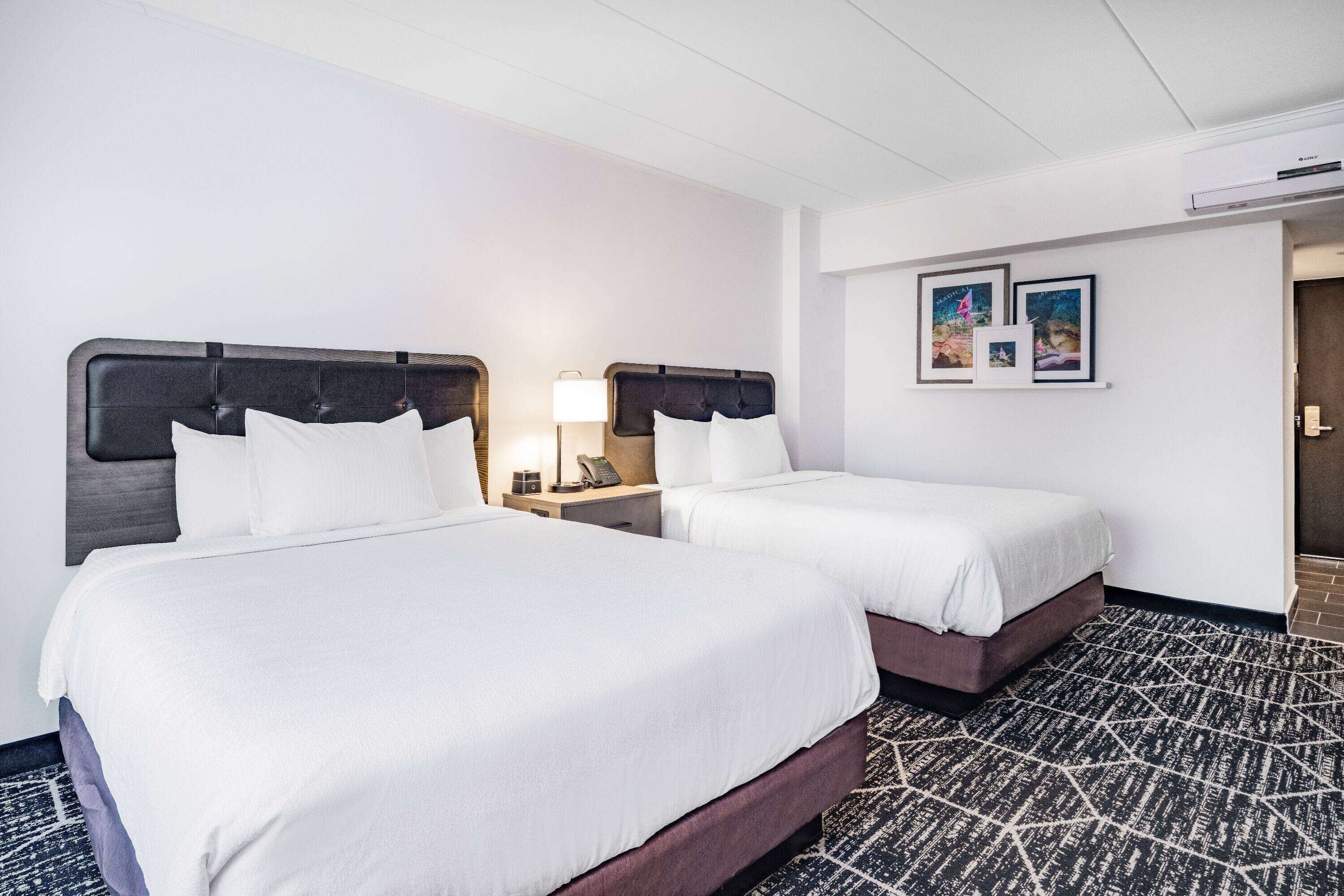 A hotel room with two beds, a nightstand with a lamp, and a wall-mounted air conditioner.