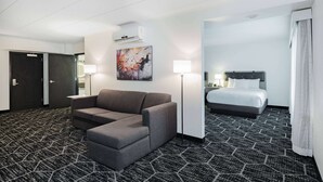 Habitación de hotel con un sofá seccional gris, una cama con ropa de cama blanca, un cuadro en la pared y una alfombra estampada.