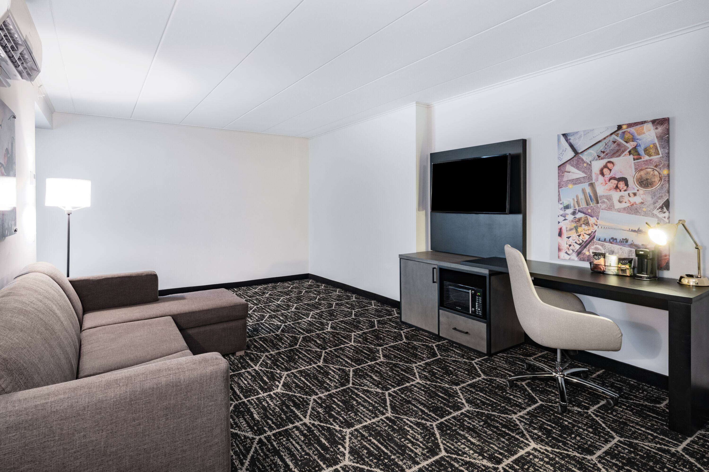 Habitación de hotel con sofá, escritorio con silla, televisor de pantalla plana, kitchenette pequeña y alfombra estampada.