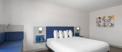 A modern hotel room with a large bed, a bedside table with a lamp, and a wall-mounted artwork.