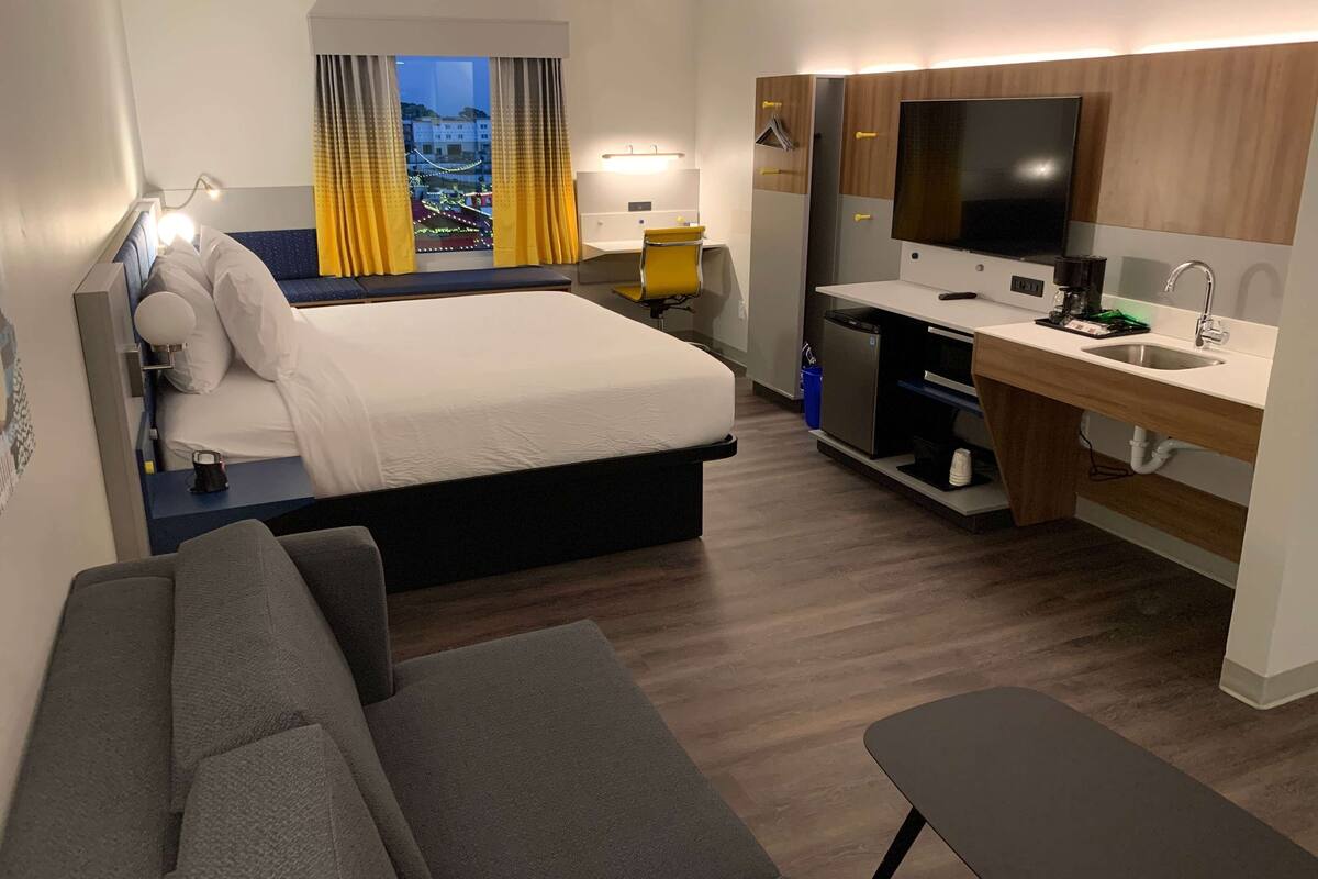A hotel room with a bed, a desk, a TV, a sink, and a view of a cityscape.