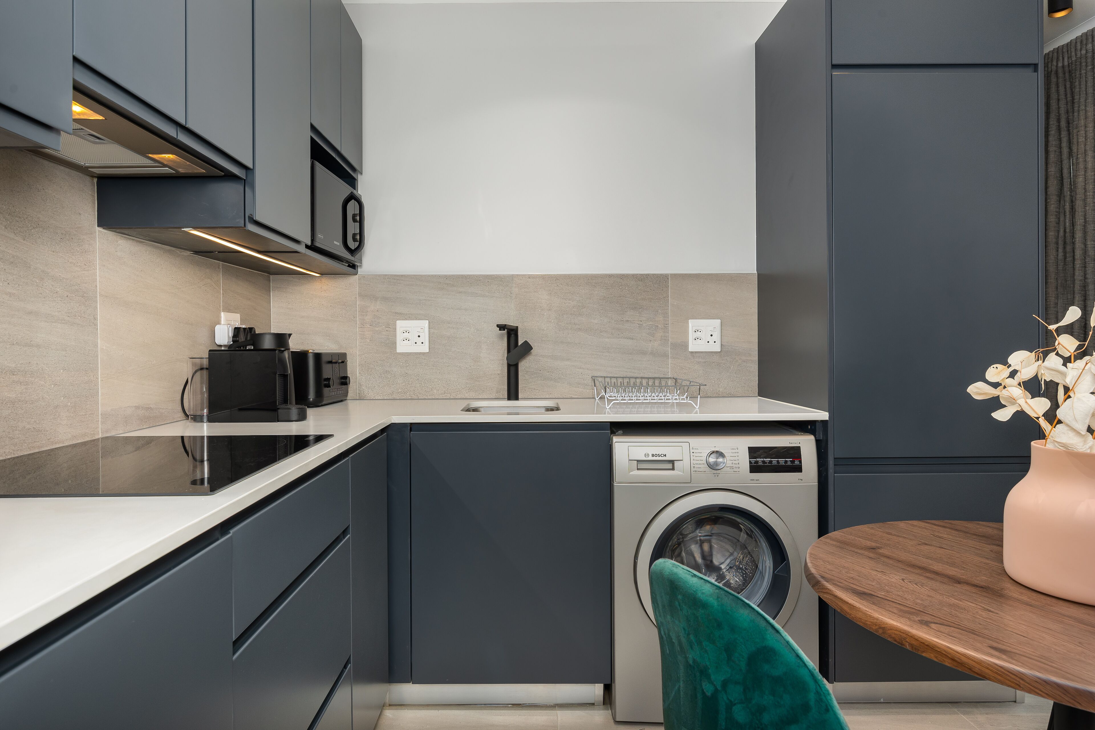 A modern kitchen with grey cabinetry, a Bosch washing machine, and a wooden table with a vase of flowers.