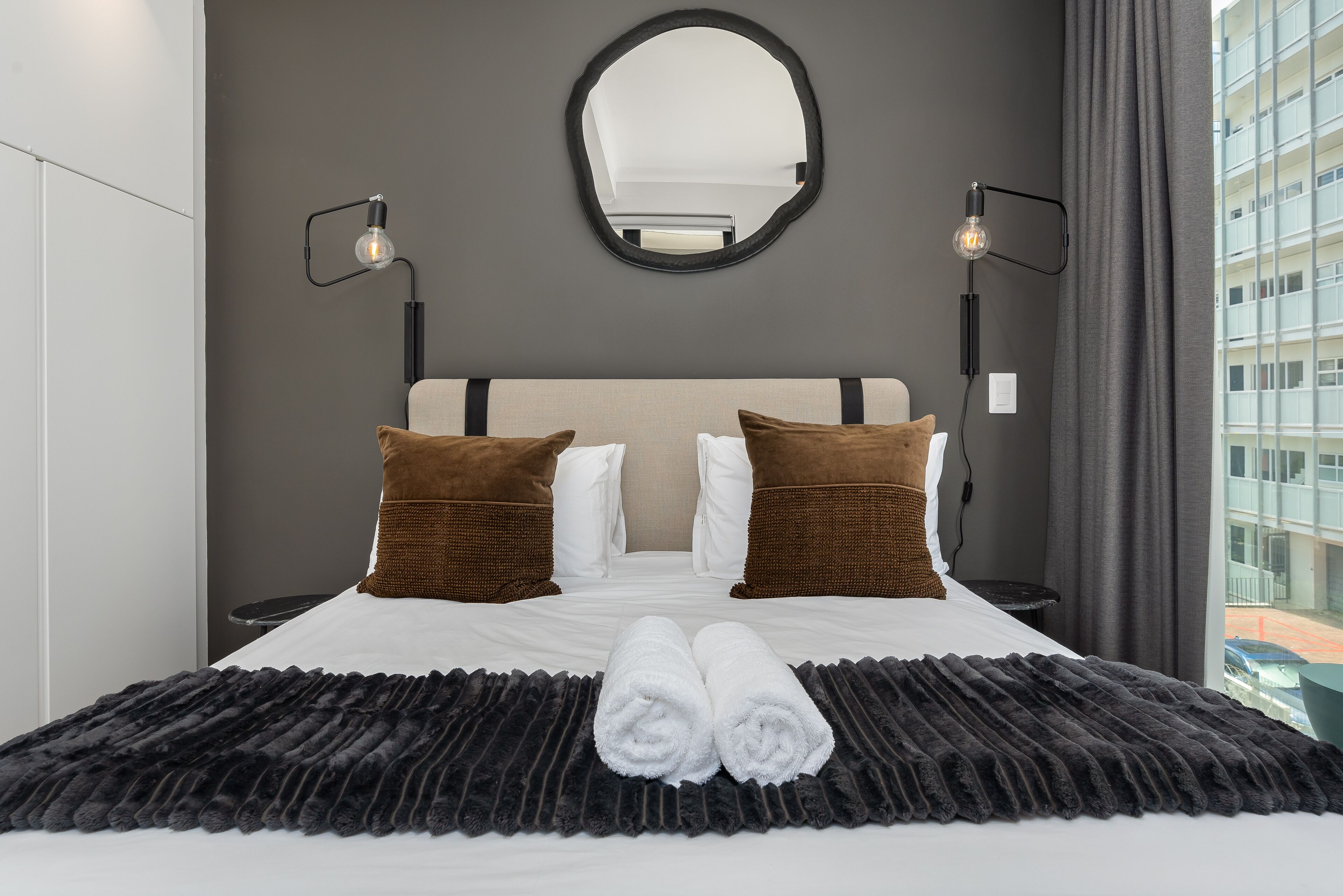 A neatly made bed with white linens, two brown pillows, and a black textured blanket. There are two wall-mounted lamps on either side of a large mirror above the bed.