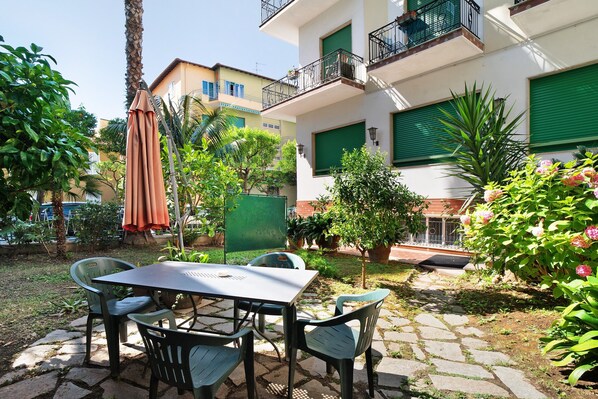 Un patio con mesa y sillas, rodeado de follaje y un edificio con balcones.