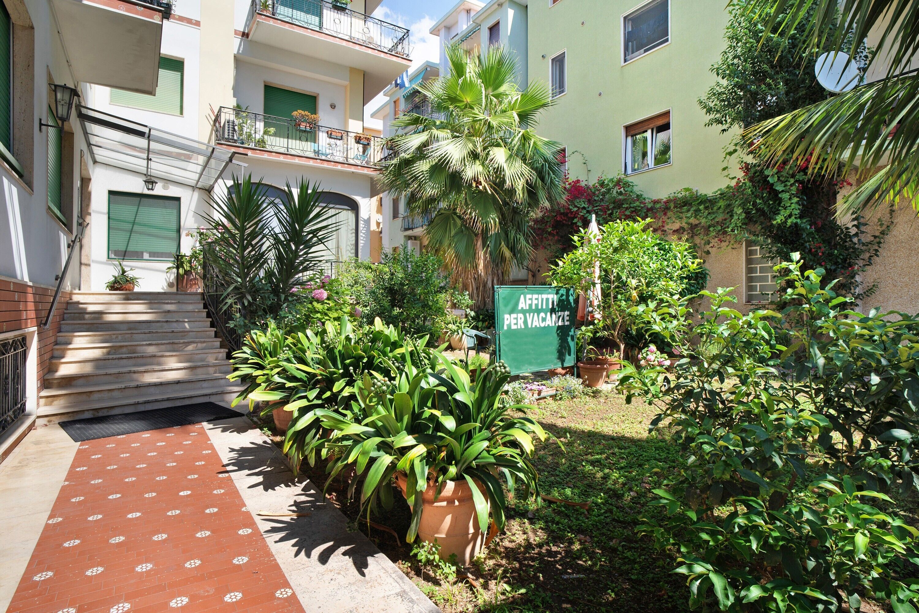 A courtyard with potted plants, a sign reading 'AFFITTI PER VACAZE,' and a staircase leading to an apartment building.