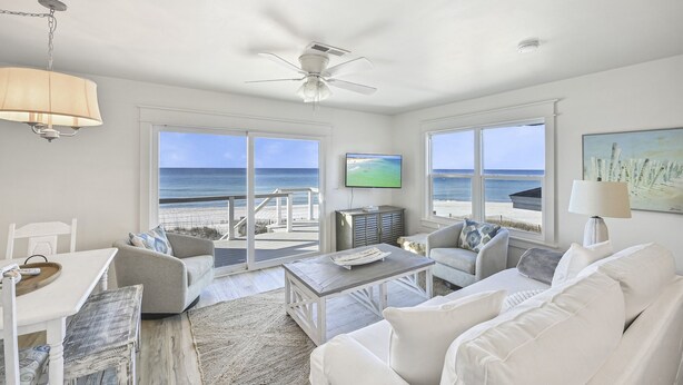 A spacious living room with a large window overlooking a beach, a ceiling fan, and a flat-screen TV mounted on the wall. - Beach Front with Balcony and Grill (Panama City Beach)