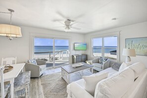 Een ruime woonkamer met een groot raam met uitzicht op een strand, een plafondventilator en een flatscreen televisie aan de muur.