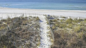 A sandy beach with a narrow path leading to the water, surrounded by sparse vegetation and a wooden fence. - Beach Front with Balcony and Grill (Panama City Beach)