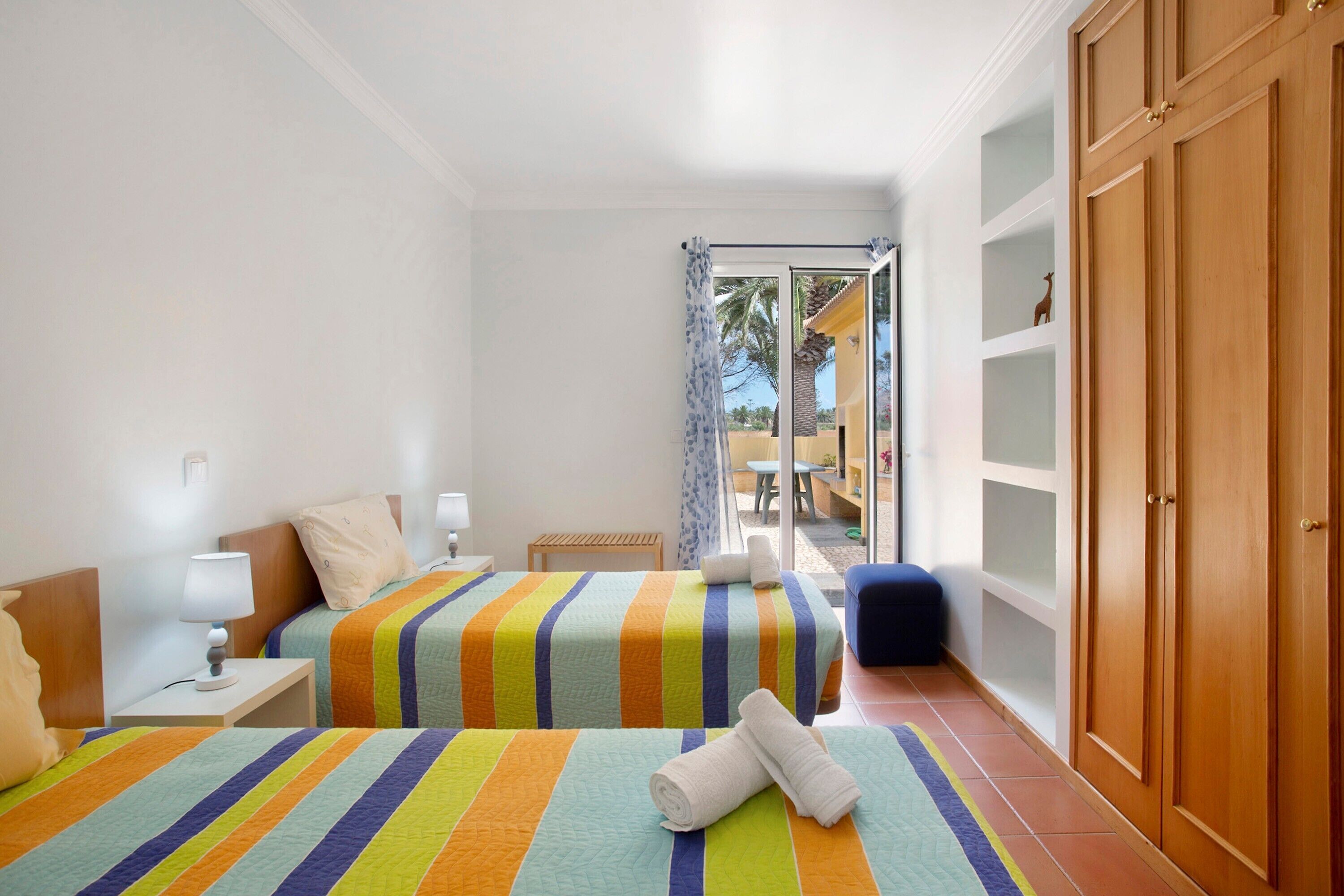 A hotel room with two beds, a wooden wardrobe, a small table with a lamp, and a view of the outside.