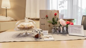 Une chambre d’hôtel avec une table en bois, un présentoir à gâteaux en verre, un vase avec des fleurs, un panneau d’accueil encadré et une lampe.