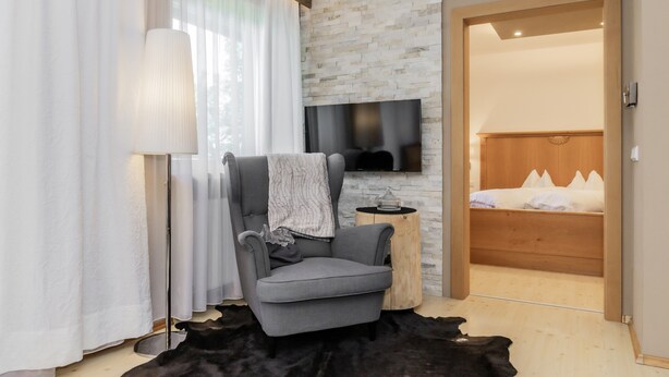 Une chambre moderne avec un fauteuil gris, un téléviseur à écran plat fixé à un mur en pierre, un lit en bois avec une literie blanche et une petite table ronde avec une lampe.