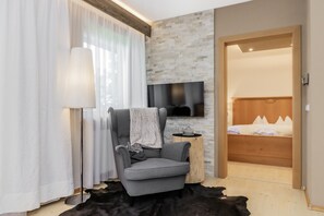 Quarto moderno com poltrona cinza, televisão de tela plana fixada em parede de pedra, cama de madeira com roupa de cama branca e pequena mesa redonda com luminária.