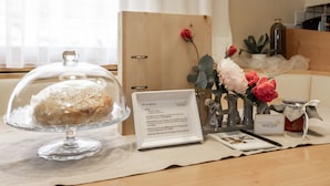 Una habitación con puerta de madera, un expositor de vidrio para pasteles, un cartel de bienvenida enmarco, una mesita con un jarrón de flores y una botella.